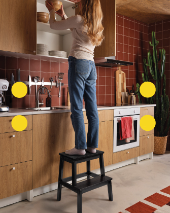 Persona de pie sobre un taburete negro alcanzando un gabinete alto en una cocina moderna.