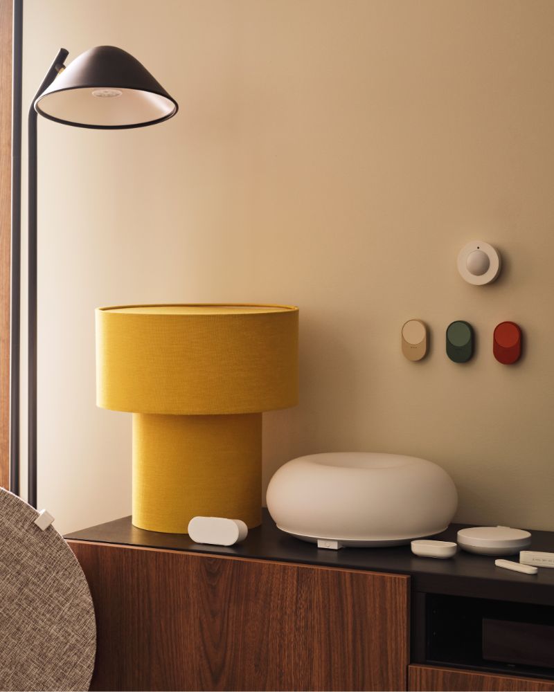 A MYGGSPRAY wireless motion sensor and three BILRESA remote controls are mounted on a wall above a TV bench next to a lamp.