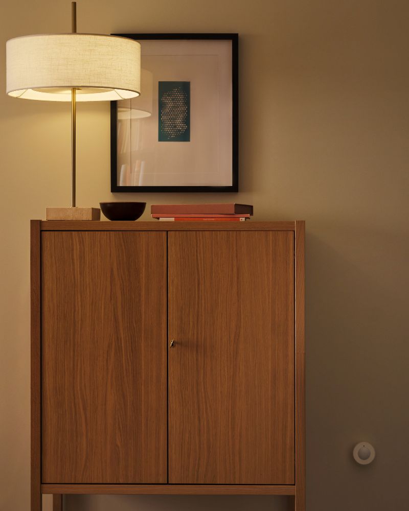 A MYGGSPRAY wireless motion sensor is mounted on a wall near a STOCKHOLM 2025 cabinet with two doors.