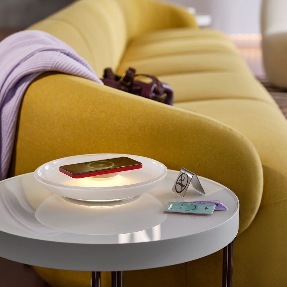 By a green-yellow sofa is a white table holding a donutshaped VÄSTMÄRKE wireless charger with lighting by hotel key cards.