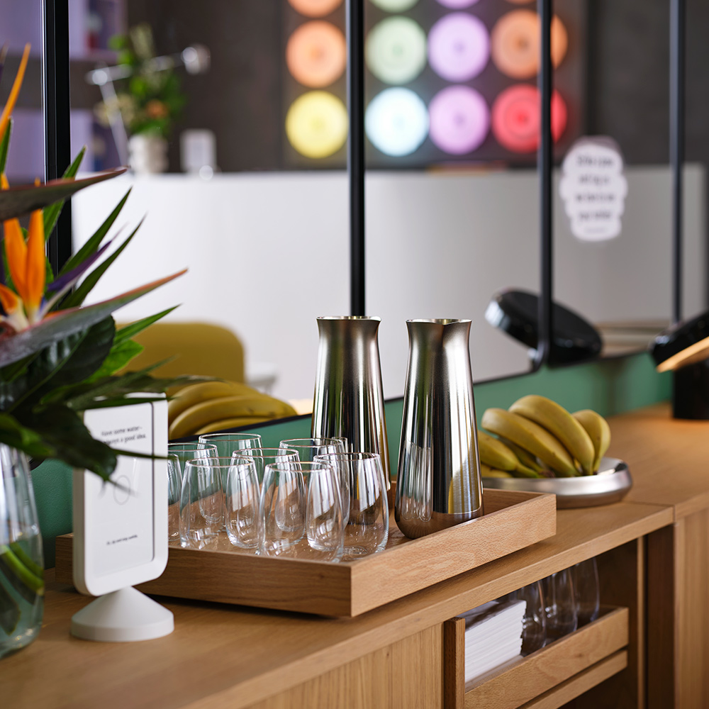Two sleek stainless steel AFTONHAJ carafes are by drinking glasses and bananas on trays as a snack station in a hotel lounge.