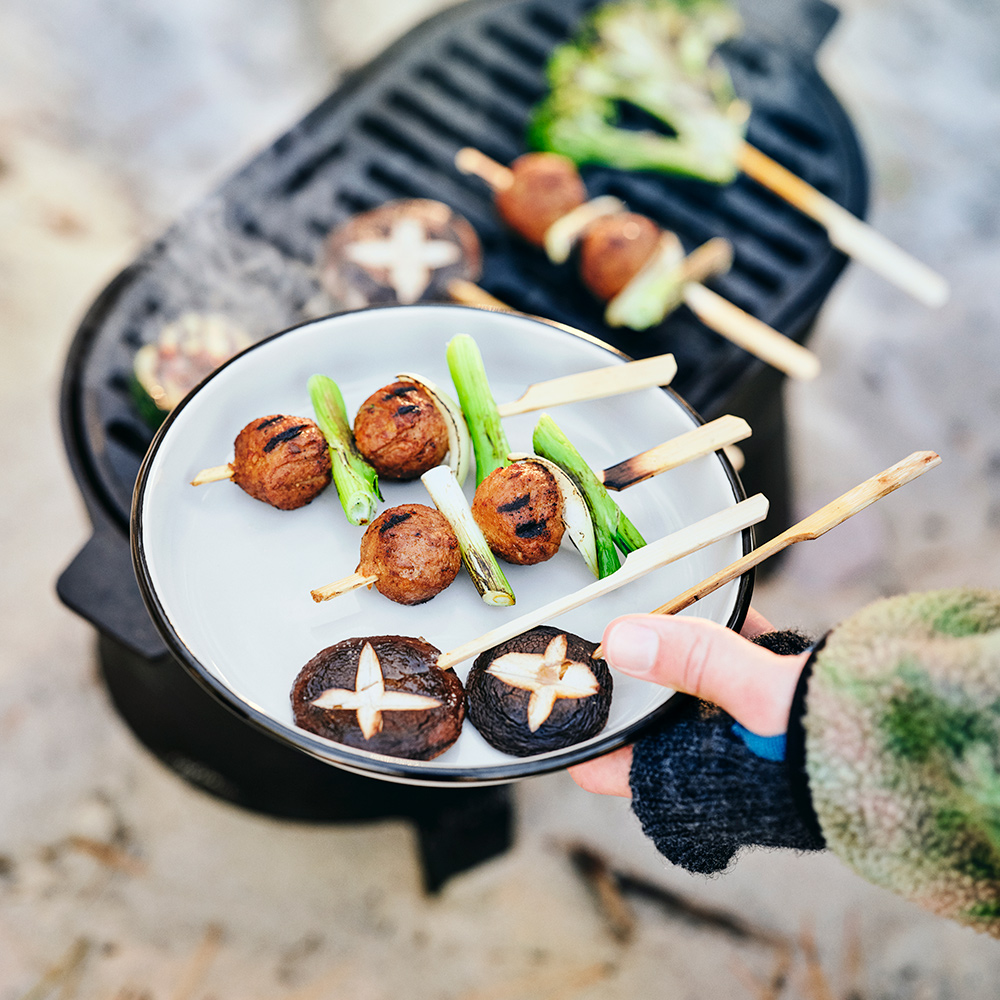 A hand holds up a SOLUPPGÅNG plate with mushroom and plantball skewers on it, close to a SOLUPPGÅNG barbecue.