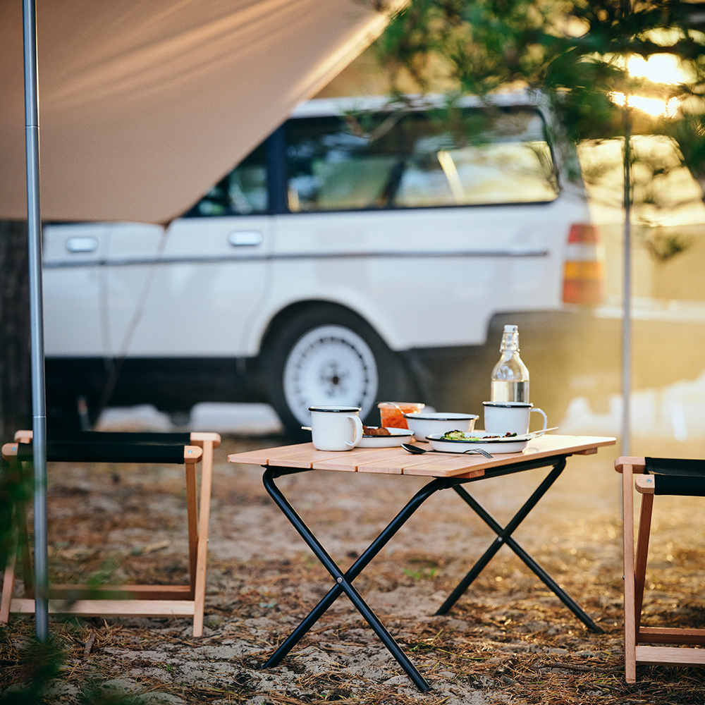 A SOLUPPGÅNG folding table with SOLUPPGÅNG plates and mugs on it is standing under a tarp, close to a car