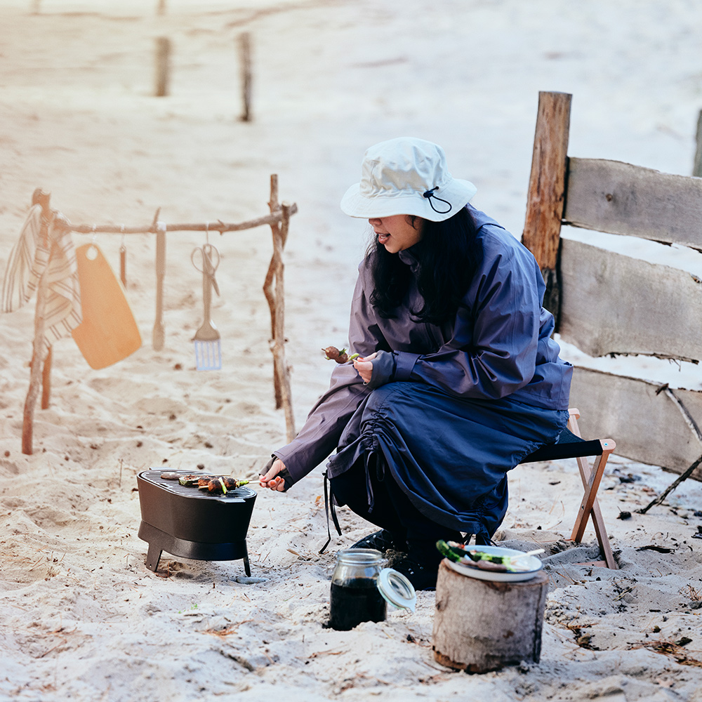 A person wearing a SOLUPPGÅNG hat is grilling skewers on a SOLUPPGÅNG barbecue, on a sandy beach.