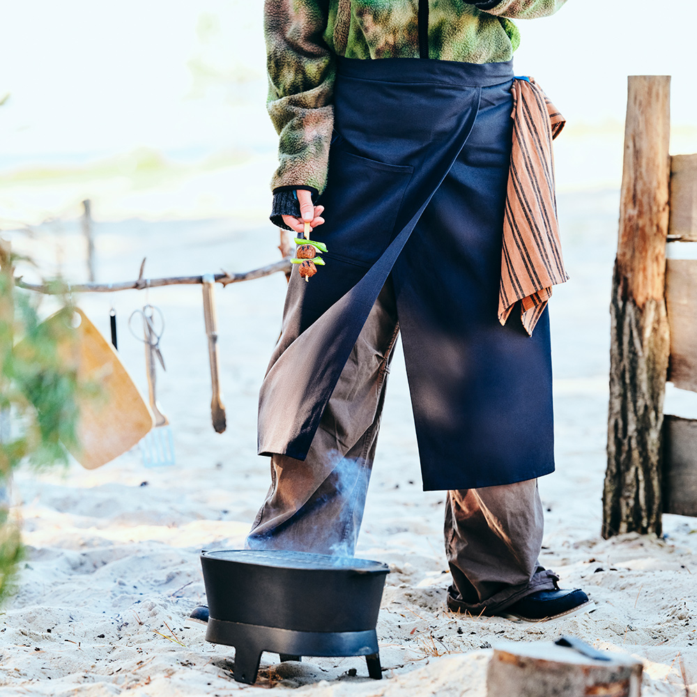 A person is wearing a SOLUPPGÅNG waist apron while standing close to a SOLUPPGÅNG barbecue, on a sandy beach.