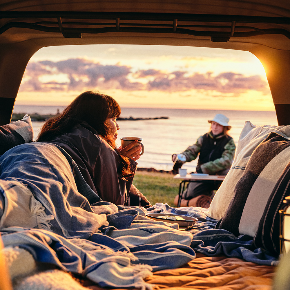 A scenic outdoor setting with a person in a SOLUPPGÅNG throw enjoying a hot drink at the back of a car, overlooking water