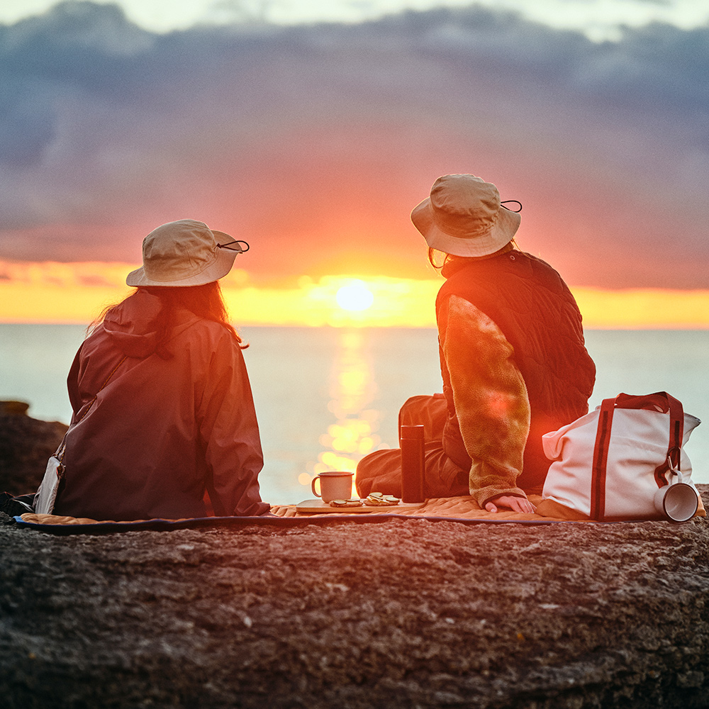 Two people wearing SOLUPPGÅNG hats are sitting on a SOLUPPGÅNG picnic blanket while watching the sunset by the sea.