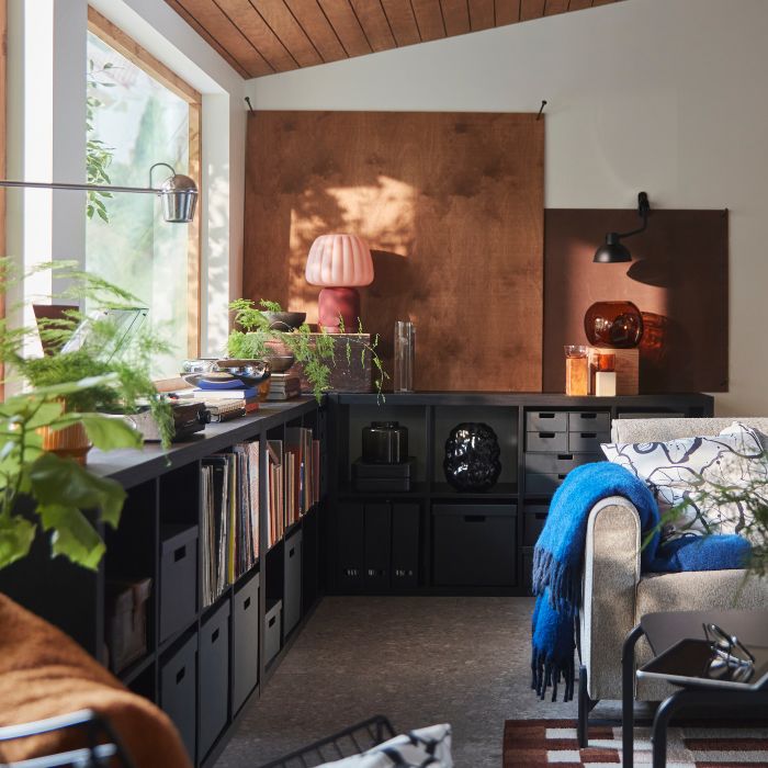 A living room features a black-brown KALLAX shelving unit holding books and decorative items beside a grey sofa.