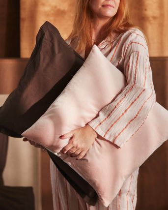 A person is holding a pillow with a dark brown HAMPFLOCKEL pillowcase and a cushion with a pale pink SANELA cushion cover.