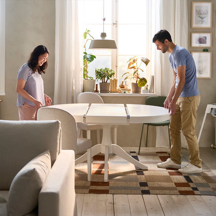 A smiling couple extends a white round dining table in a bright, cozy room with plants and framed art.
