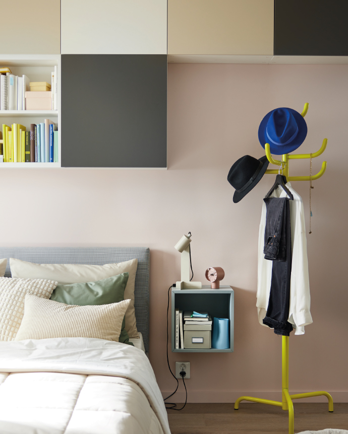 Modern bedroom with a white bed, modular wall shelves, a floating bedside table with a lamp and clock, and a yellow coat rack holding clothes and hats.