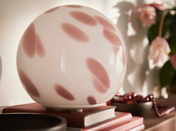 A close-up of a pink FADO table lamp made of glass that sits on stacked books beside a brown bowl and a plant.