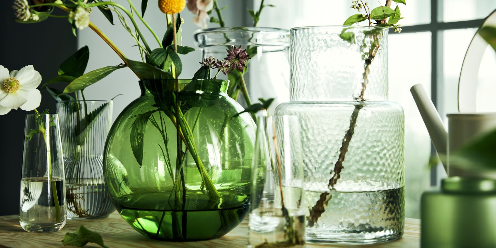 Table with vases, featuring PÅDRAG, KONSTFULL, BEGÄRLIG and BERÄKNA glass vases arranged for a bright floral display.