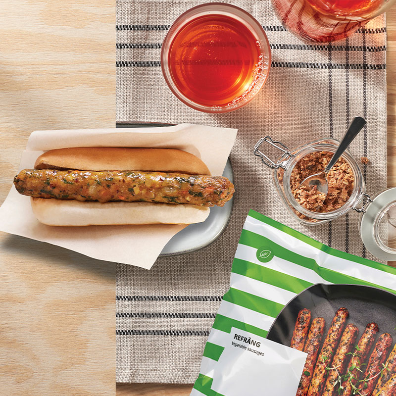 A vegetable sausage in a bun sits on a plate next to a packet of REFRÄNG sausages, a glass of juice, and a jar of crispy onions on a wooden table.