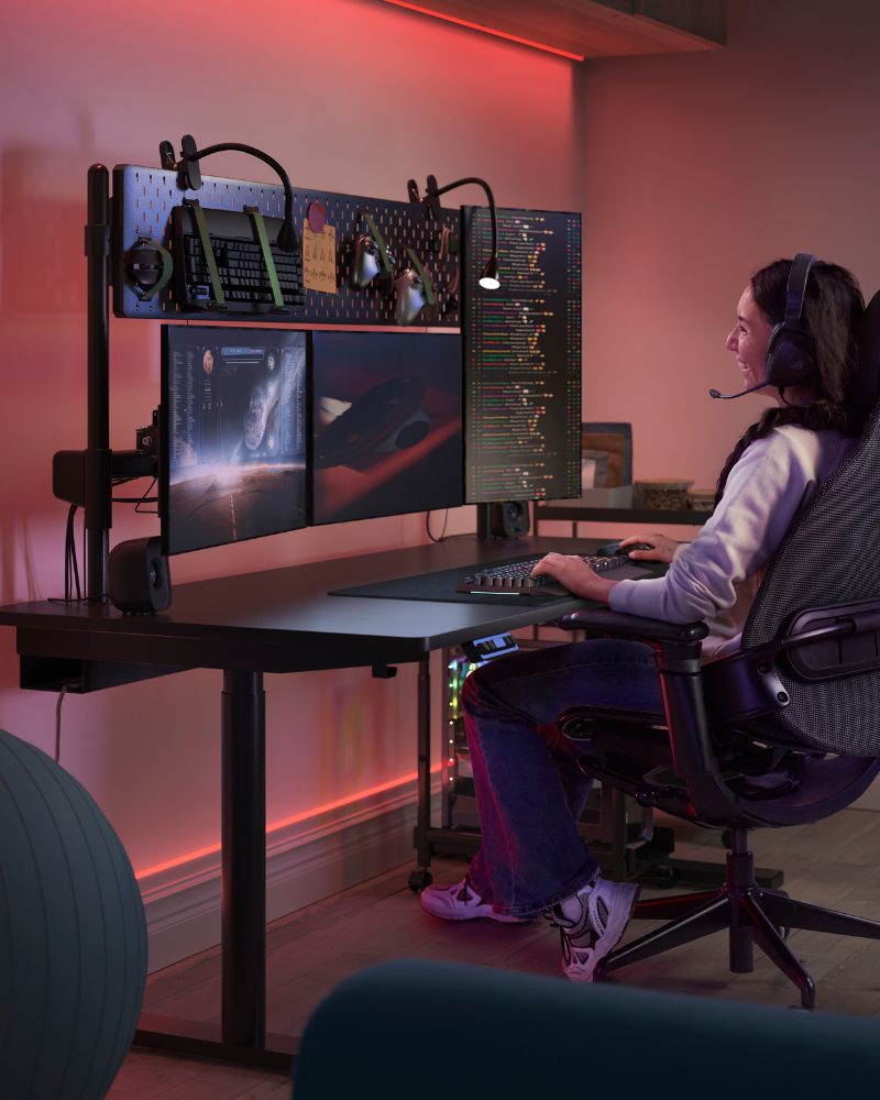 A gaming setup with MÅLOMRÅDE sit/stand desk and BÄSTBOLL chair, lit by NÄVLINGE clamp lights in a vibrant, immersive space.