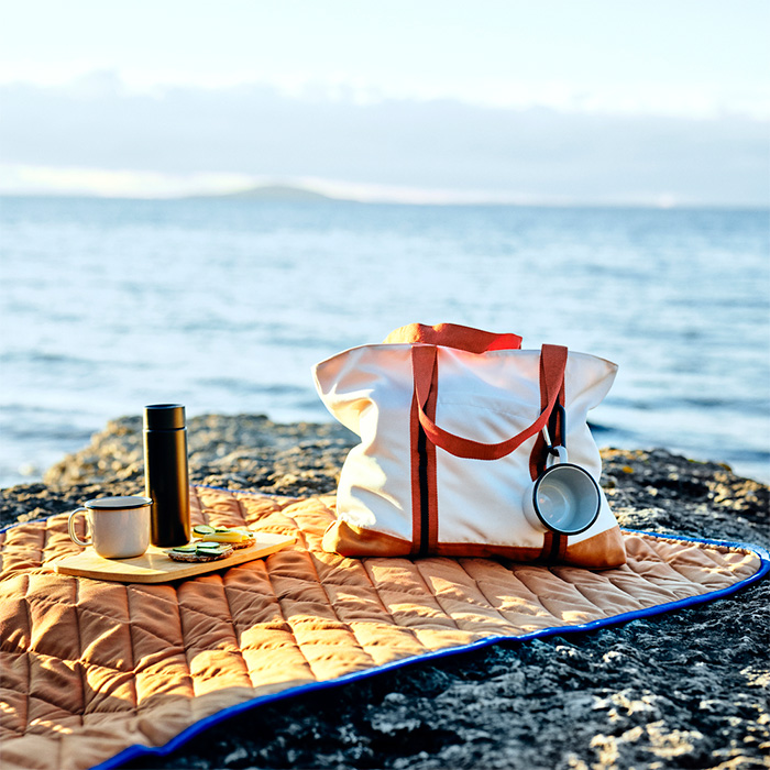 Escena de picnic costero con bolso, manta acolchada y accesorios sobre rocas frente al mar.