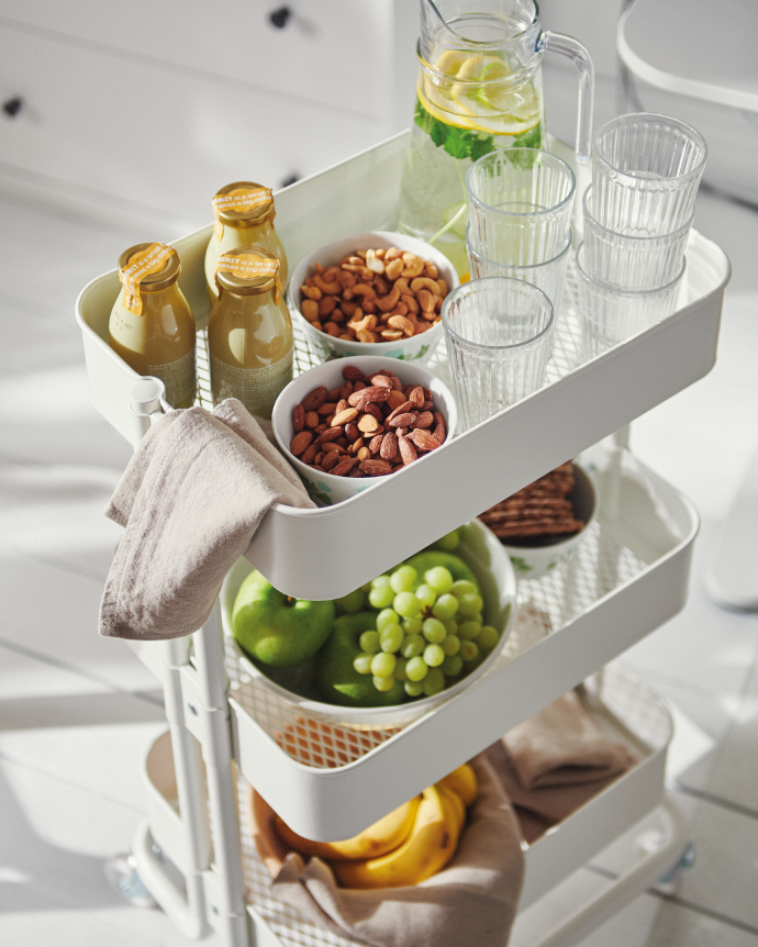 White three-tier storage cart with drinks, glasses, nuts, and fresh fruits like grapes, apples, and bananas in a bright kitchen.