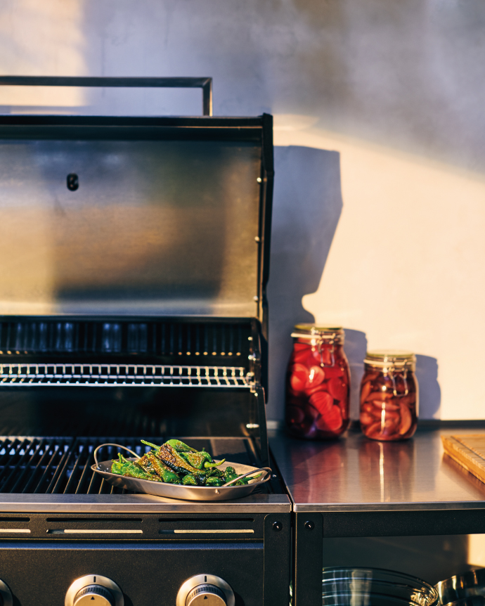 Open stainless steel grill with a pan of roasted green peppers, next to jars of pickled vegetables on a countertop.