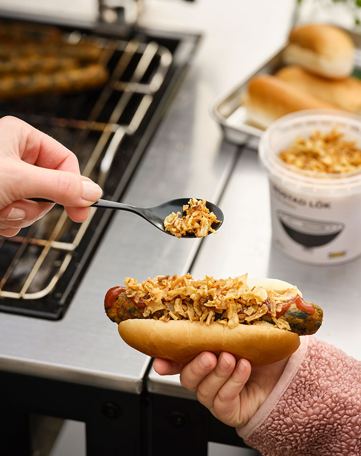 A child’s hand holding a vegetable hot dog in a bun while an adult adds crispy onions on top with a black spoon near a grill.