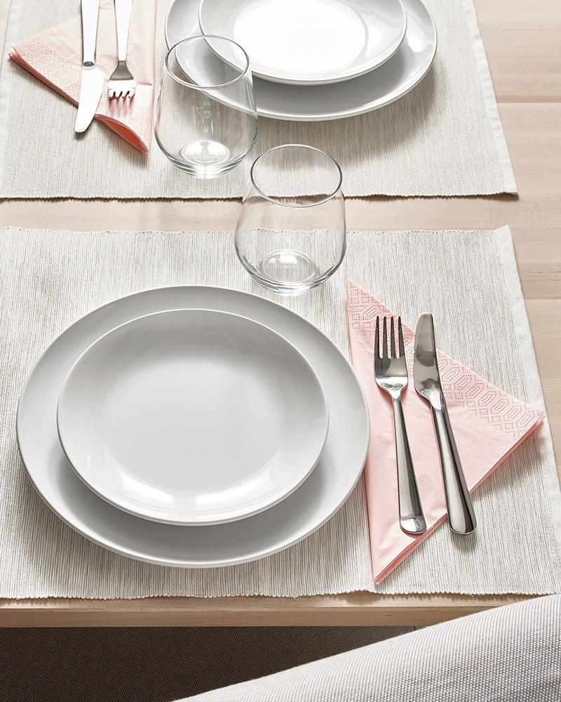 A GODMIDDAG plate on top of a tinted GODMIDDAG plate, next to a glass cup and a DRAGON fork‑and‑knife set on a dining table.