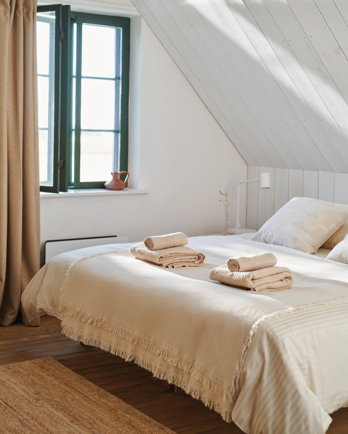 Bright minimalist bedroom with a slanted ceiling, beige bedding, folded towels on the bed, and a green-framed window next to cream-colored curtains.