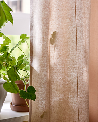 Cortina beige con textura junto a una planta verde en el alféizar de una ventana con suaves sombras de hojas.