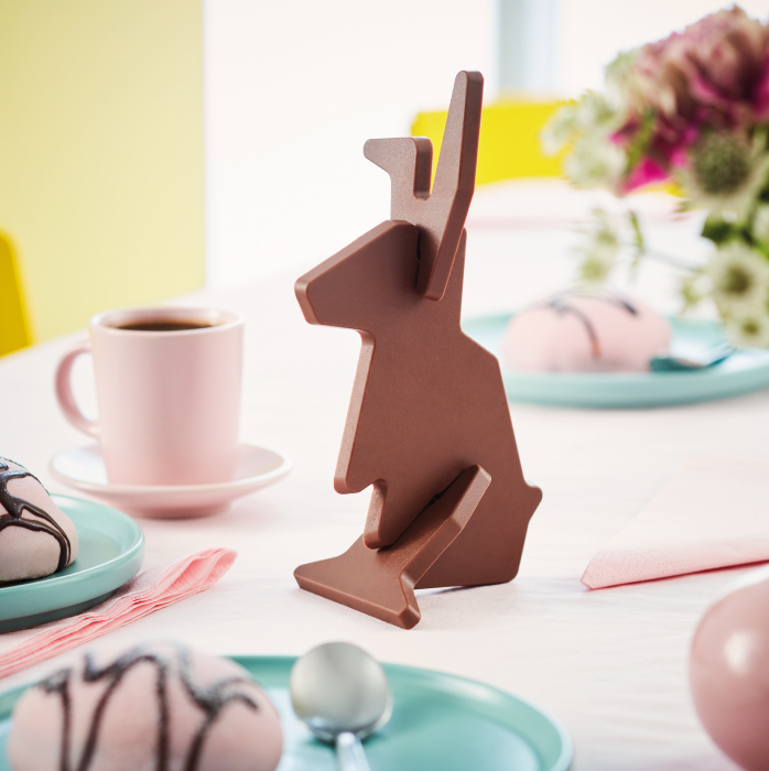 Chocolate bunny-shaped figure standing on a pastel table set with coffee and desserts.