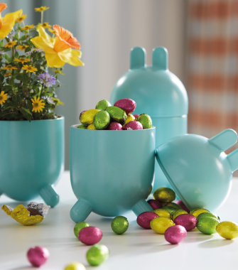 Turquoise bunny-shaped containers filled with colorful foil-wrapped chocolate eggs next to a small flower arrangement.