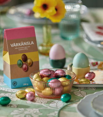 A festive table setting featuring a paper bag labeled "VÅRKÄNSLA," a ribbed bowl filled with multi-colored foil chocolate eggs, two pastel eggs in cups, and yellow flowers in a vase, all on a patterned green tablecloth.