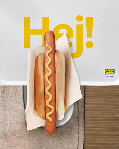 A top-down photograph of an IKEA hot dog with mustard on a bun, placed on a white tray with "Hej!" text and the IKEA logo, next to a cloth napkin on a wooden table.
