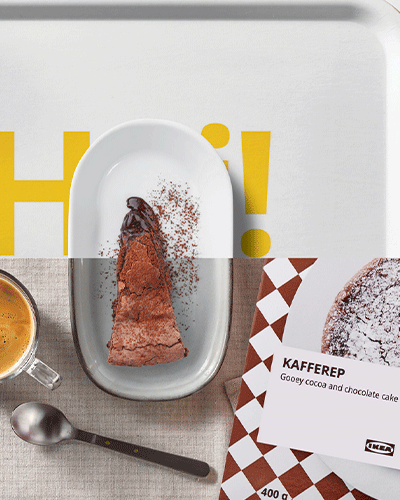 A top-down photograph of a slice of chocolate cake on a plate, a cup of coffee, a spoon, and an IKEA KAFFEREP package on a cloth napkin. The background shows part of a white tray with "H!" text and the IKEA logo.
