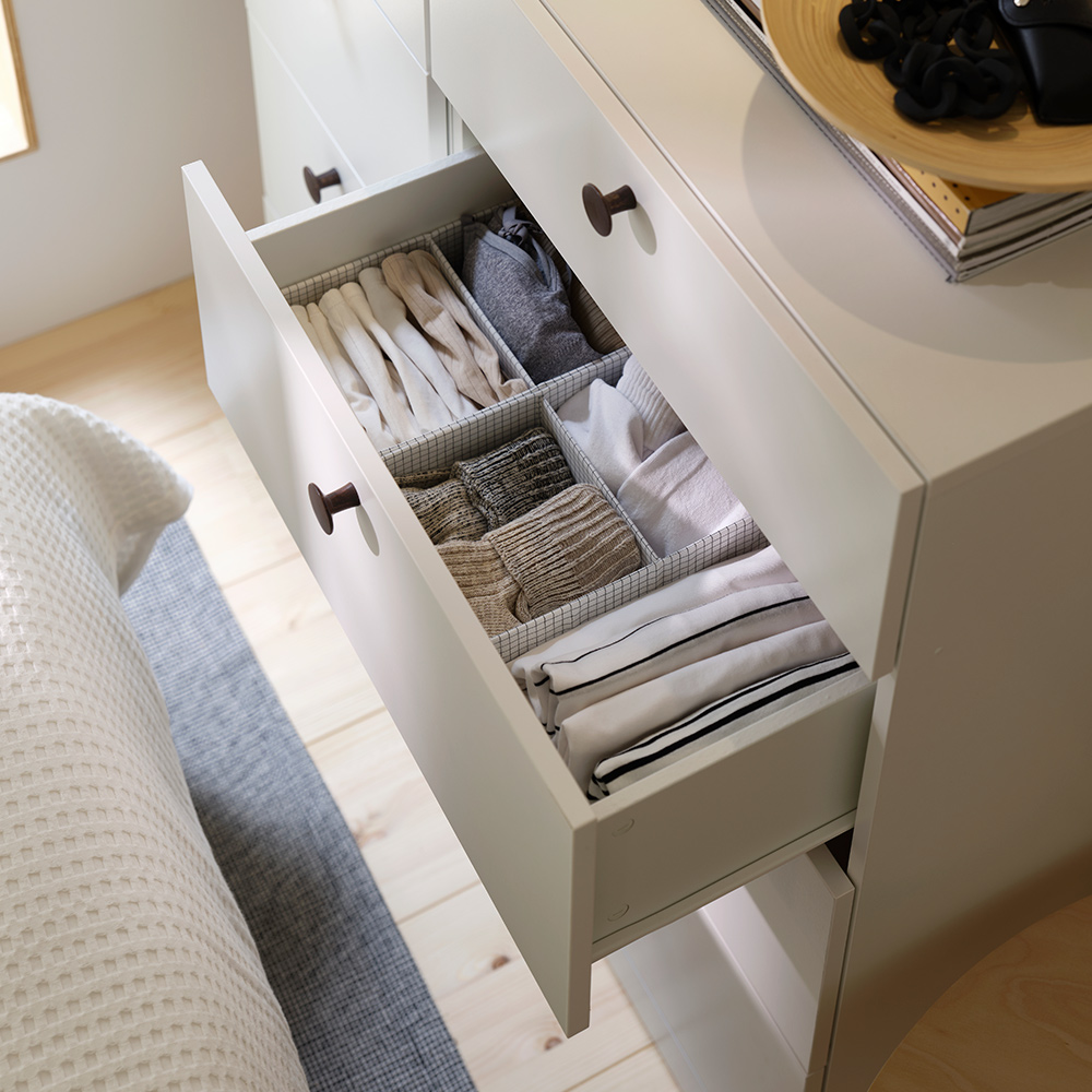 An open drawer revealing a STUK box with compartments organising folded clothes inside a white chest of drawers.