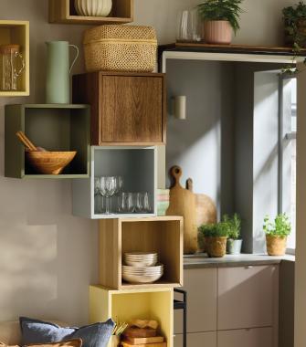 Estantería modular en la pared con cubos de diferentes colores y madera que contienen platos, vasos, plantas y utensilios de cocina.