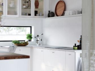 Cocina moderna blanca con gabinetes superiores con vitrina, encimera clara, plantas y utensilios de cocina junto a una ventana.