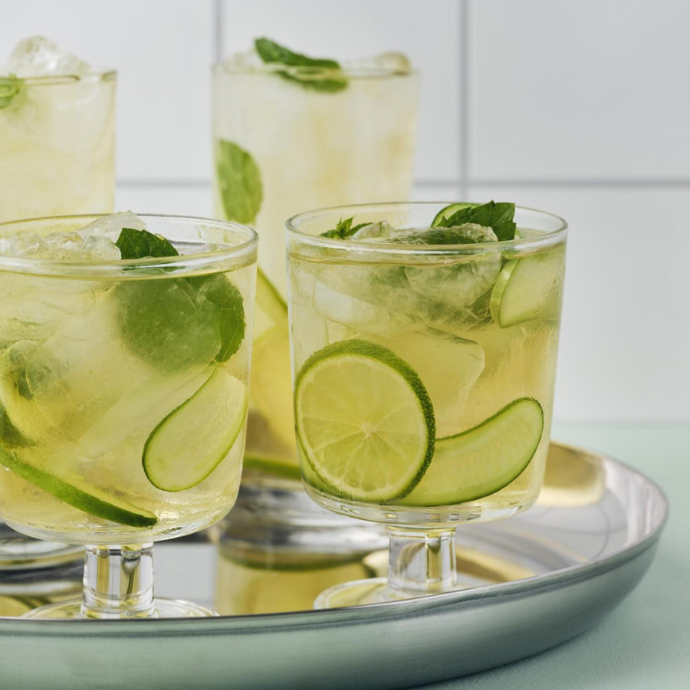 Three glasses of chilled drink with ice, lime slices, cucumber, and mint leaves on a metal tray.