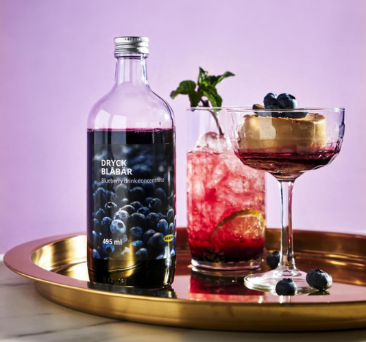 Bottle of blueberry drink concentrate next to two iced cocktails with blueberries and mint on a golden tray.