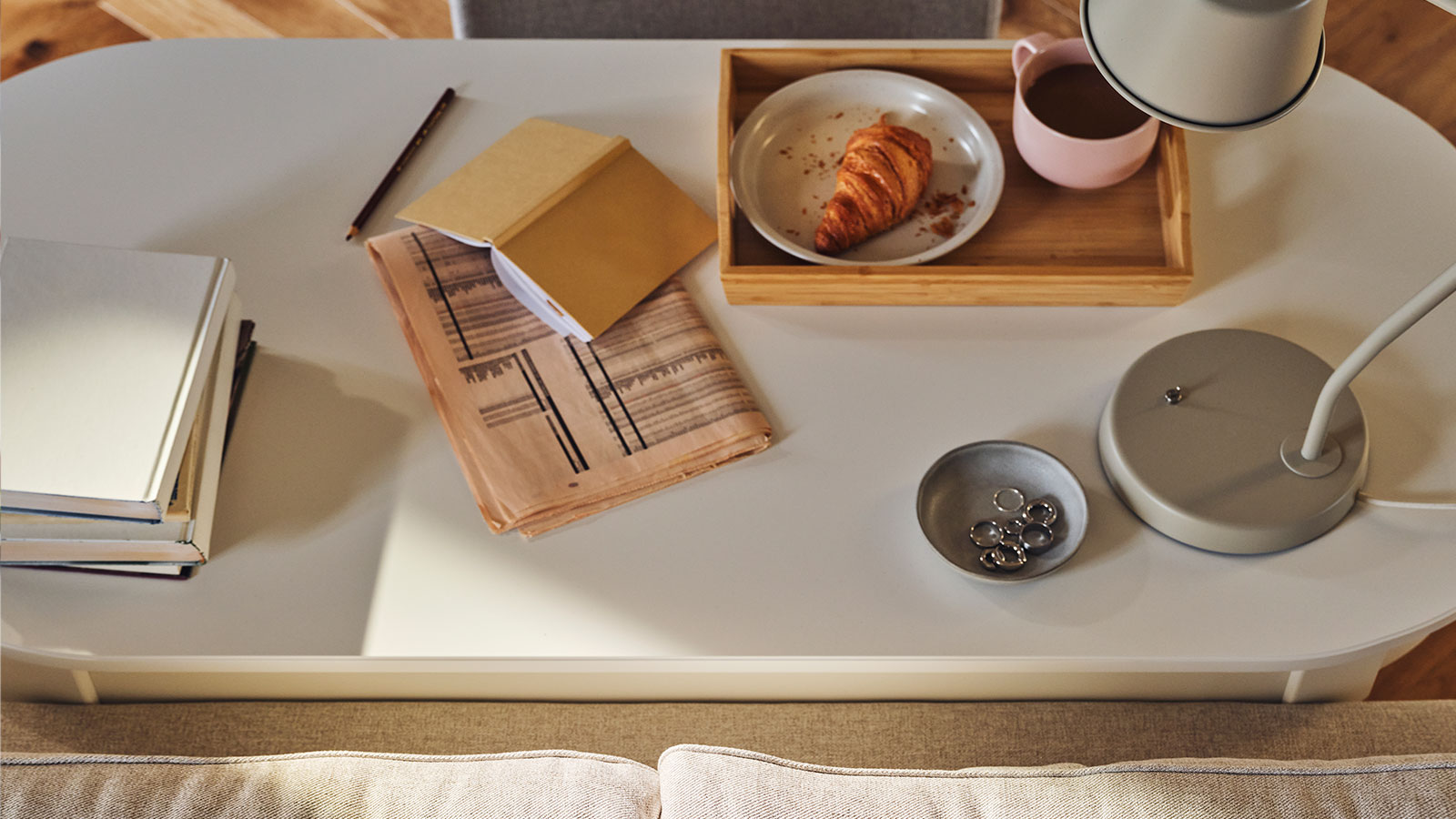 A tray with food and coffee, a lamp, a book and a newspaper stand on a ROSENTORP desk behind an EKHOLMA 2-seat sofa bed.