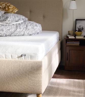 Cama tapizada beige con colchón blanco y ropa de cama estampada junto a una mesita de noche de madera con lámpara y decoración.