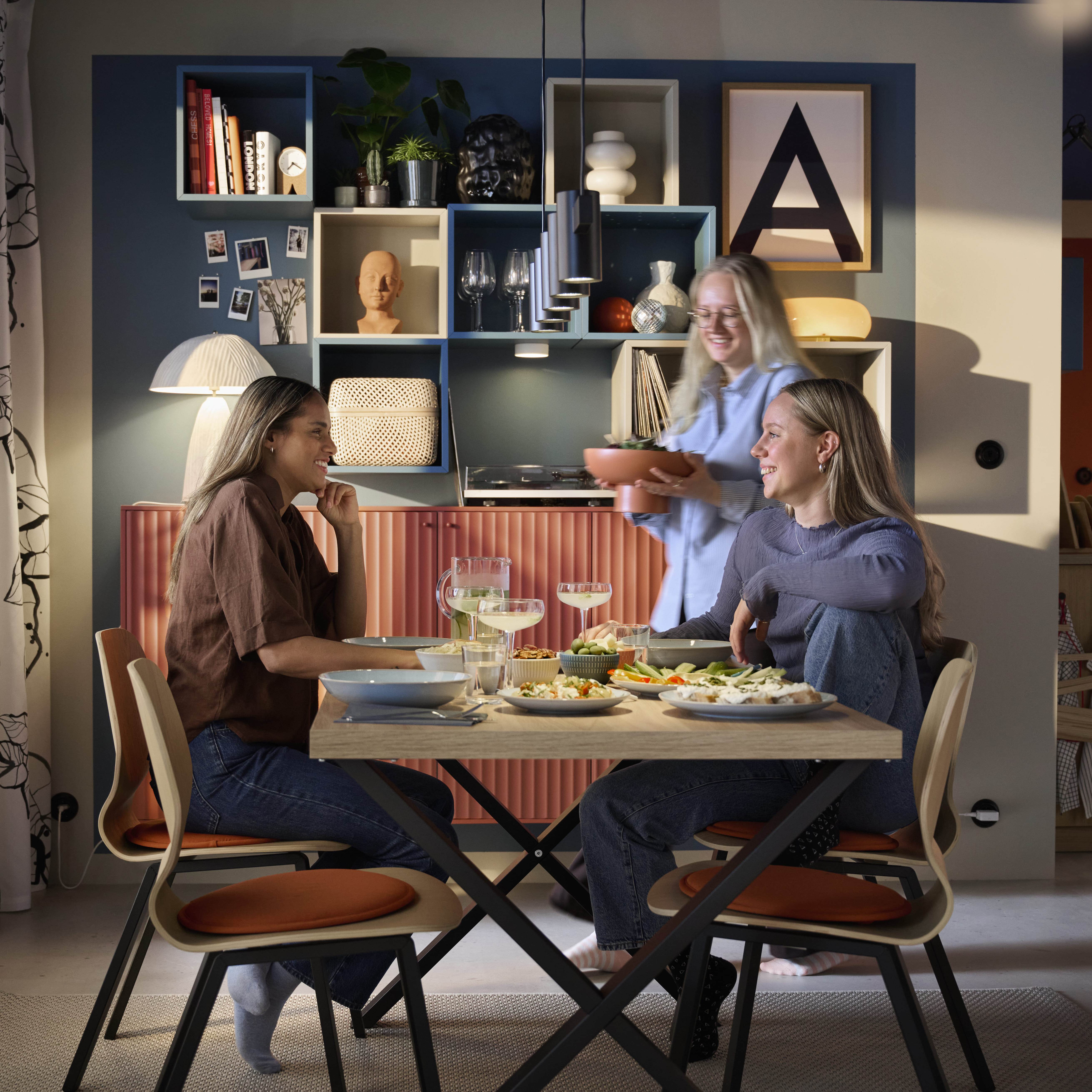 Tres amigas cenando en un comedor moderno con estanterías coloridas y una iluminación cálida y acogedora.