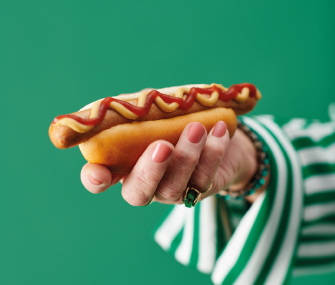 Mano sosteniendo un hot dog con ketchup y mostaza sobre fondo verde.