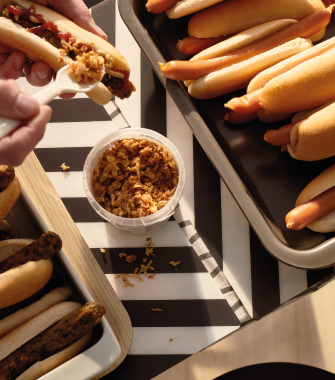 Bandejas con hot dogs y un recipiente con cebolla frita; una mano agrega topping a un pan.