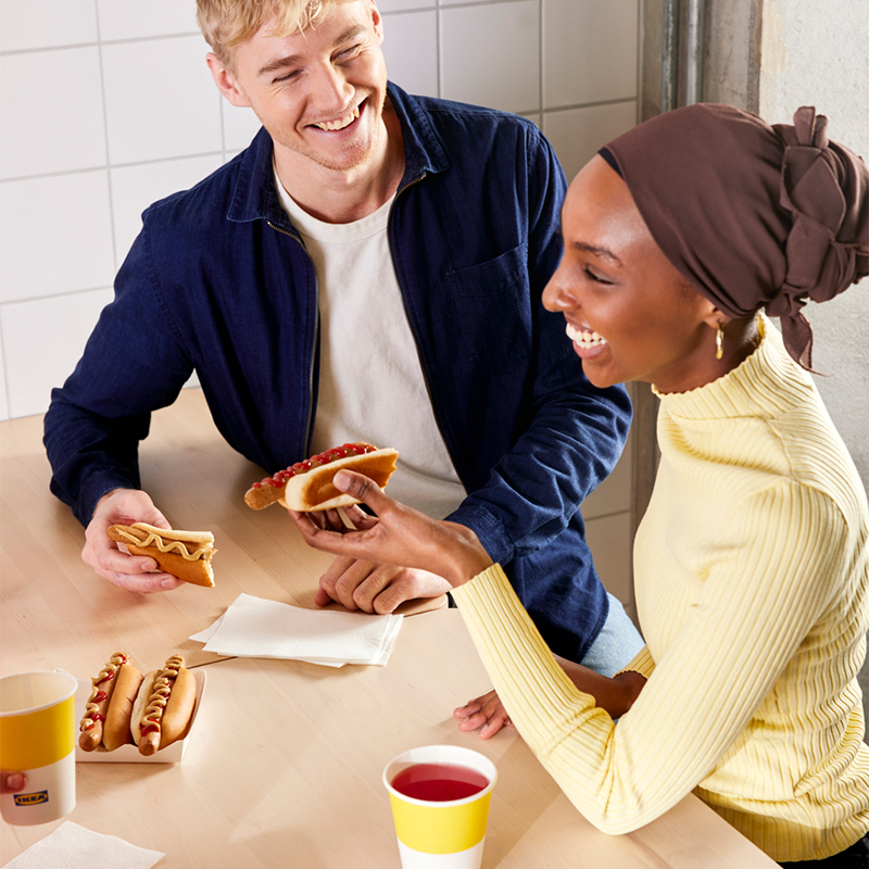 A man and a woman in a hijab laugh at a wooden table while enjoying hot dogs and drinks in a bright setting.