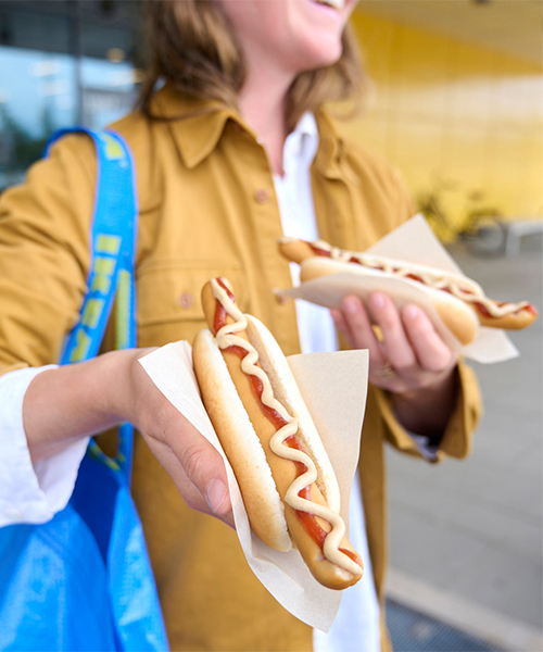 Close-up of a person in a mustard jacket with a blue IKEA bag holding two hot dogs with condiments.