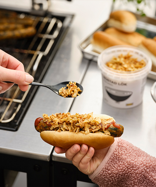 Close-up of a person spooning fried onions onto a veggie hot dog held by someone in a pink fleece coat next to a grill.