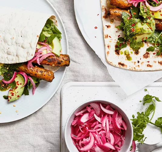 Top-down view of two flatbread wraps filled with veggie sausages, avocado, pickled onions, and green sauce, next to a bowl of extra onions.