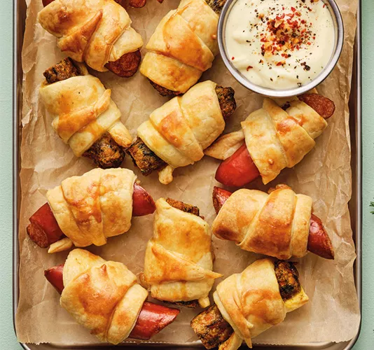 Top-down view of several golden pastry-wrapped sausage rolls on parchment paper, served with a small bowl of white dipping sauce topped with spices.