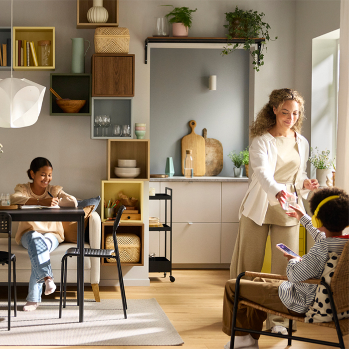 A bright home scene featuring a woman handing a glass of water to a child wearing headphones, while a young girl studies at a nearby table. The space is decorated with organized colorful cube shelving and indoor plants.
