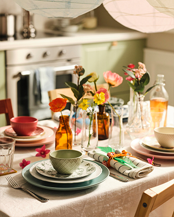 Dining table set with colorful dinnerware, fresh flowers in glass vases, and linen napkins in a cozy kitchen setting.