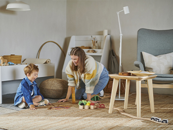 A woman and a young child play with a wooden train set on a natural fiber rug in a bright playroom.