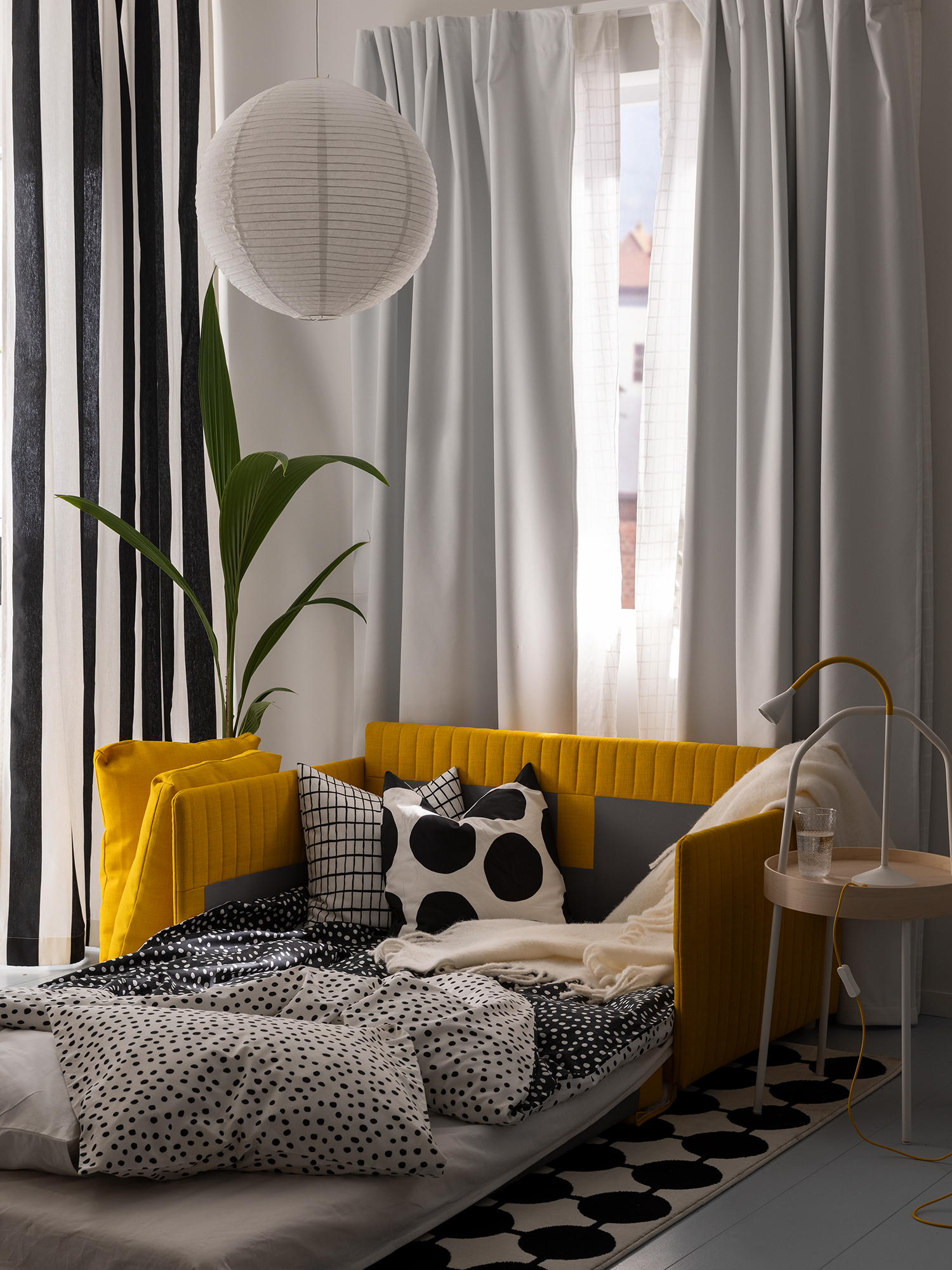 Dormitorio acogedor con sofá cama amarillo, cojines estampados en blanco y negro, cortinas claras y una planta junto a la ventana.